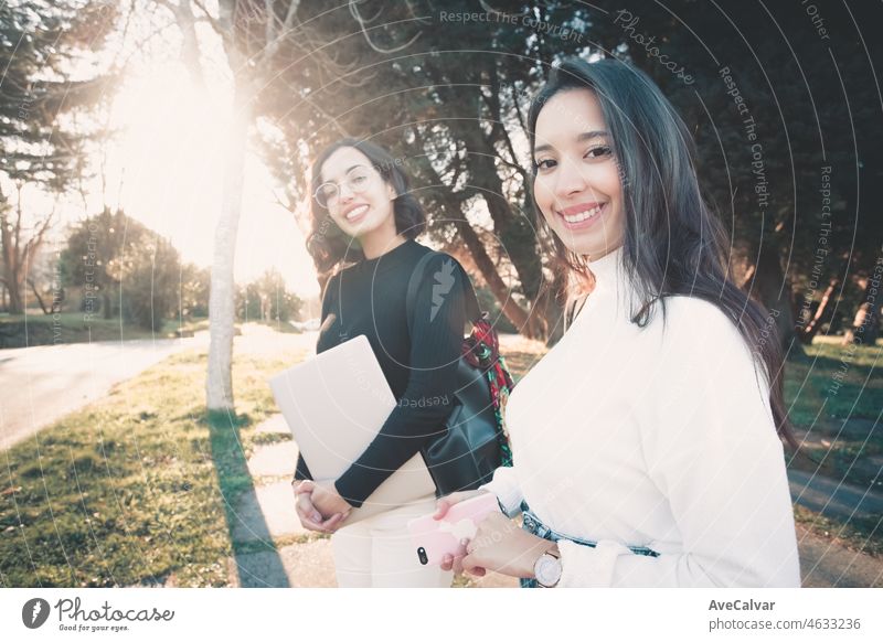 Two young students standing outdoors holding laptop, backpack and study accessories while waiting for the bus to come and take them home. Studying outside home concept. Back to campus