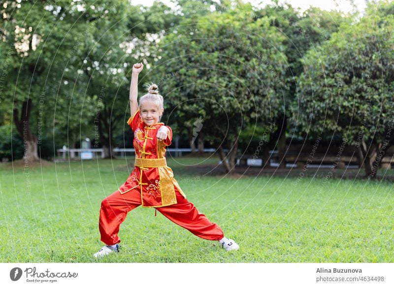 Cute little caucasian girl seven years old in red sport wushu uniform exercising in park at summer day. Lifestyle portrait of kung fu fighter child athlete