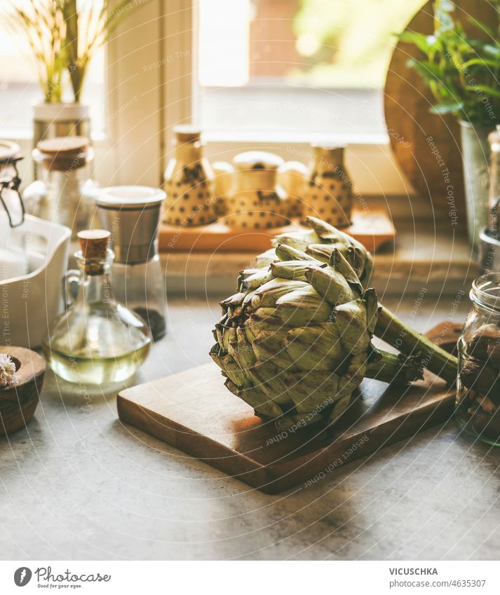 Close-up of whole artichoke on cutting board on kitchen table with ingredients close-up kitchen utensils window background natural light cooking home