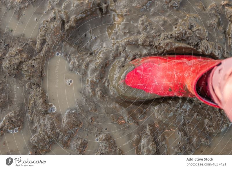 Anonymous kid standing on dirty ground pathway gumboots mud puddle wet water street childhood rubber sidewalk color walkway bright footwear surface road leg red