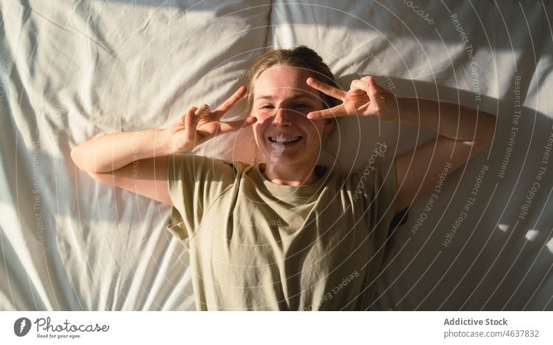 Joyful young lady lying on bed and looking at camera woman smile comfort cheerful relax bedroom arm raised holiday home joy cozy female casual rest happy