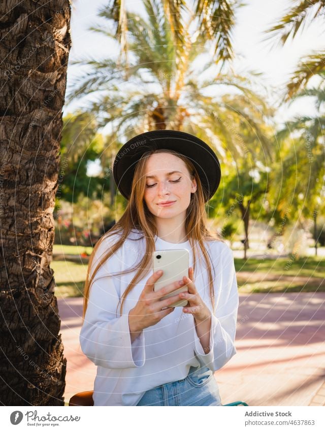 Young cyclist using smartphone in park woman social media summer palm lean female young tree flower rest style hat daytime casual browsing mobile lifestyle