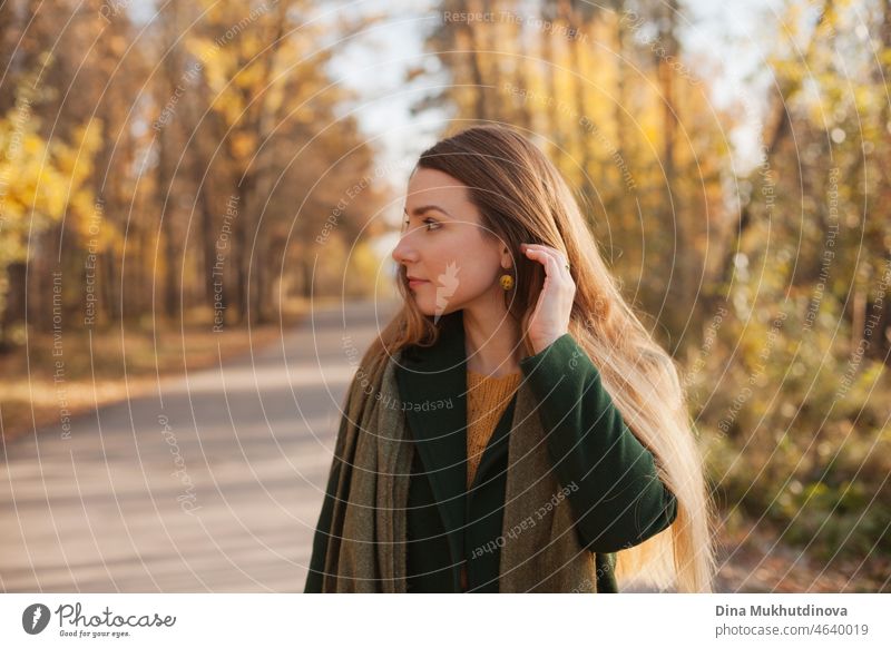 Beautiful woman in green coat and yellow sweater in autumn park near trees with yellow leaves at sunset. Spending time in nature to keep peace in mind, relaxing moments and minfullness. Autumn style and fashion, casual clothing.