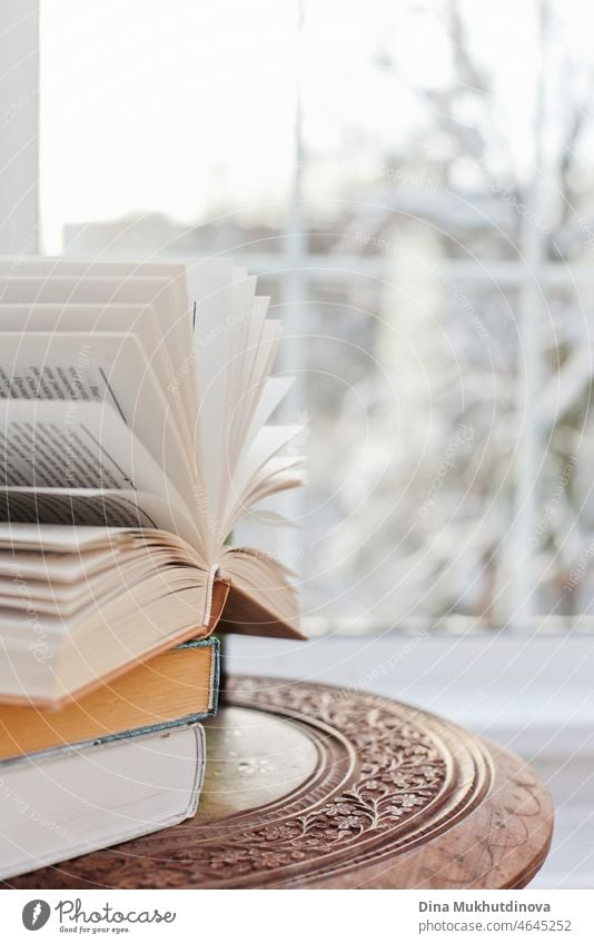 A stack of books on the wooden coffee table near armchair in the room with snowy trees landscape window view. Cozy home in winter. Winter mood Winter vacation