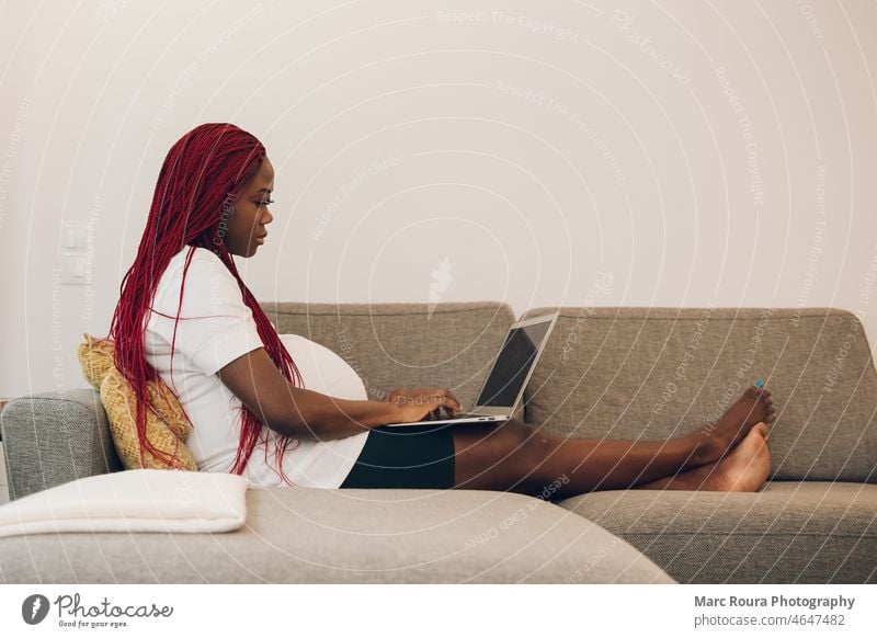 Black pregnant woman relaxing on sofa at home with his laptop abdomen adult anticipation baby belly birth business care chinese computer couch expecting family