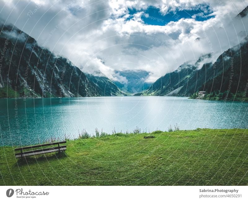 A wooden bench in front of the Schlegeisspeicher in the Zillertal in summer with clouds and sunshine in the mix Zillertaler Alps ice cream storage schlegeis