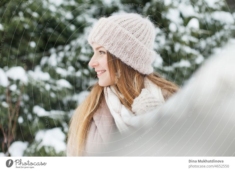 Beautiful caucasian woman with long hair walking in winter forest with snow on her knitted beanie hat. Winter fashion and stylish outfit. Real people having fun in winter, enjoying fresh air in nature with snowy fir trees.