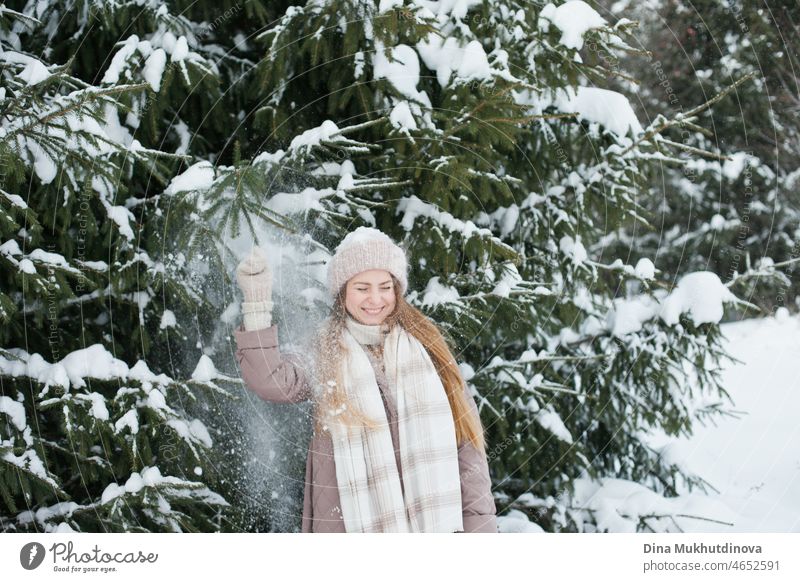 Beautiful caucasian woman with long hair walking in winter forest with snow on her knitted beanie hat. Winter fashion and stylish outfit. Real people having fun in winter, enjoying fresh air in nature with snowy fir trees.