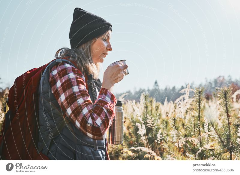 Woman taking break and relaxing with cup of coffee during summer trip. Woman standing on trail and looking away. Woman with backpack hiking through tall grass along path in mountains