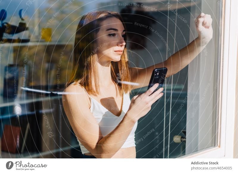 Young woman with smartphone near window using mobile internet thoughtful connection online browsing female young social media surfing communicate pensive