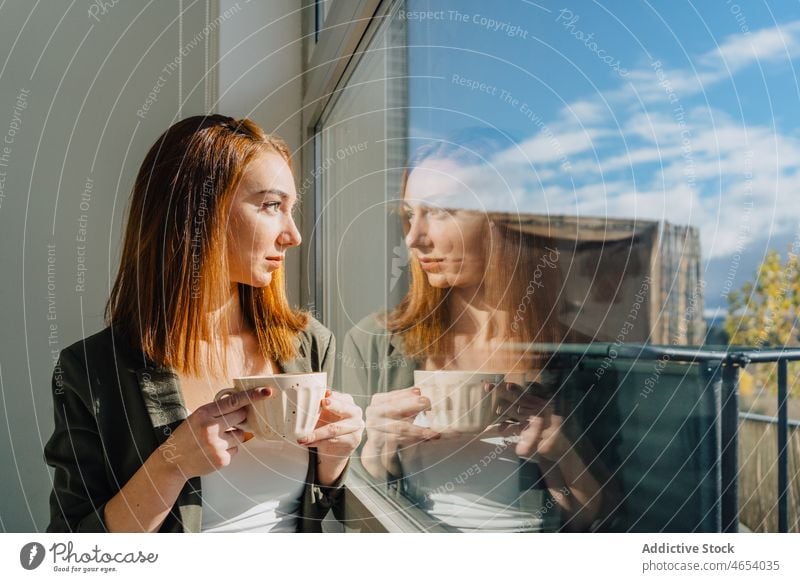 Woman standing neat windows with cup of coffee woman drink relax dreamy comfort warm rest serene female windowsill tea cozy lounge ginger red hair redhead enjoy