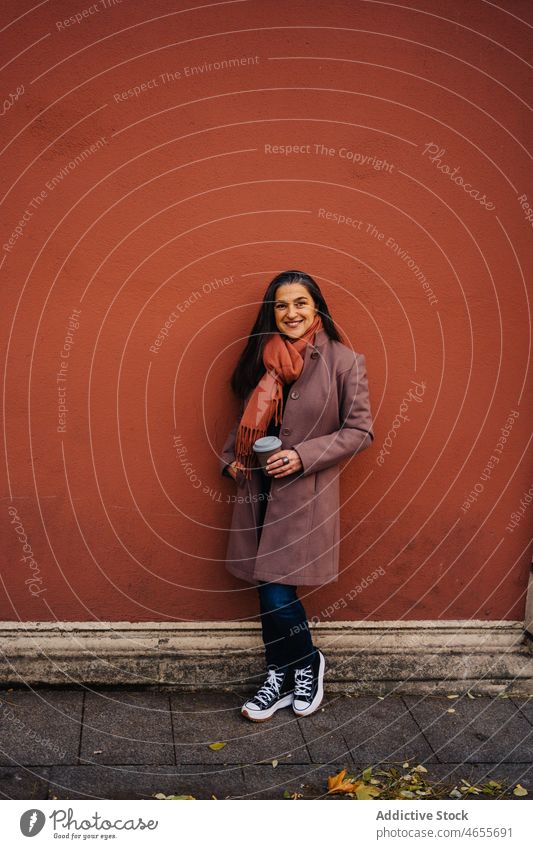 Hispanic lady with coffee cup against wall woman takeaway smile to go cheerful beverage happy sidewalk urban adult mature pavement street female stand hot drink