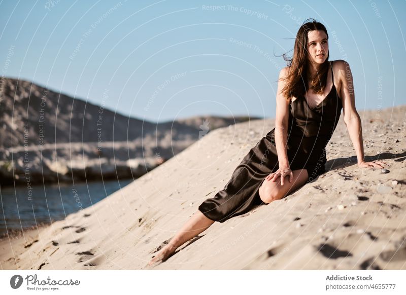 Slim attractive woman sitting on sandy beach in sunny day desert summer cloudless seashore blue sky barefoot seaside dress harmony sundress coast travel rest