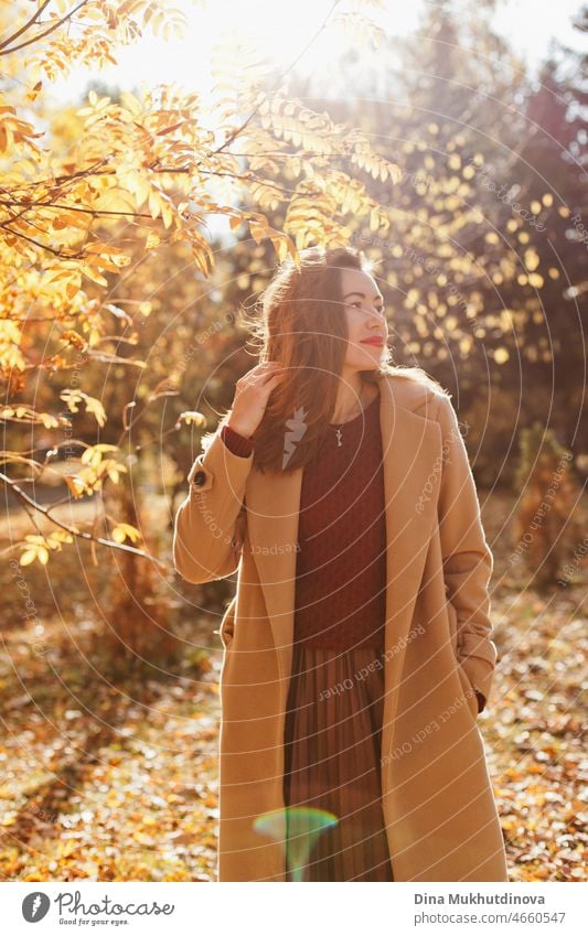 Beautiful woman in beige coat, burgundy sweater, brown skirt and red lipstick make up, standing in autumn park. Portrait of a girl in autumn nature with yellow foliage.