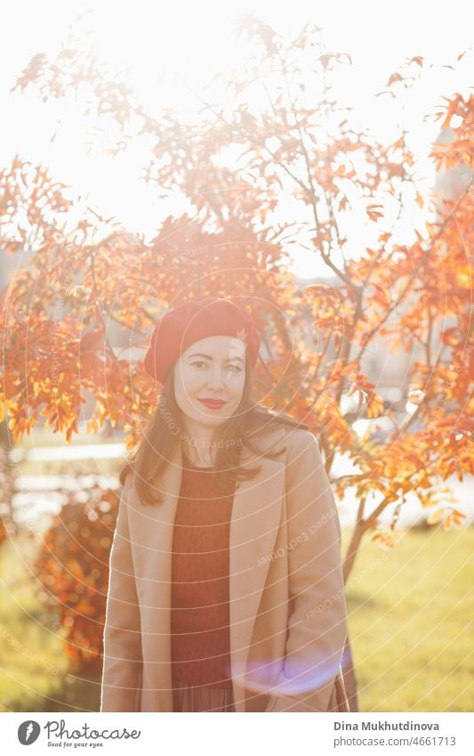 Young woman in red beret, beige coat and burgundy sweater and red lipstick make up, standing near autumn tree. Portrait of a girl in autumn park with orange red foliage.