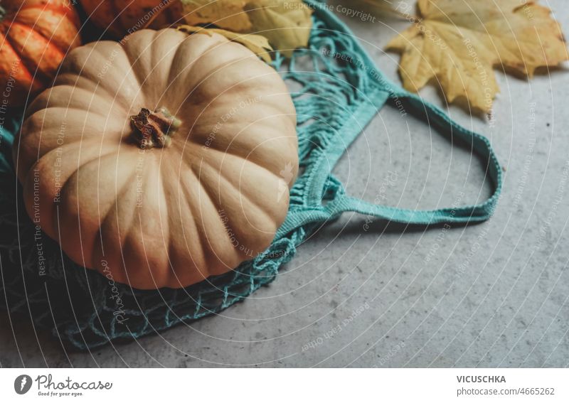 Pumpkin in blue reusable shopping bag with yellow autumn leaves. pumpkin sustainable lifestyle eco friendly zero waste grey concrete kitchen table utensils