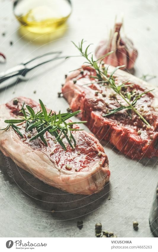 Close up of raw beef steak with green pepper and rosemary at grey kitchen table close up butcher fork olive oil preparing fresh meat barbecue top view