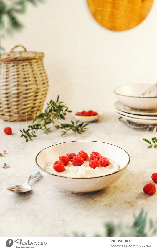 Breakfast porridge bowl with raspberries breakfast white kitchen table spoon plates utensils branches wall background still life front view berry breakfast idea