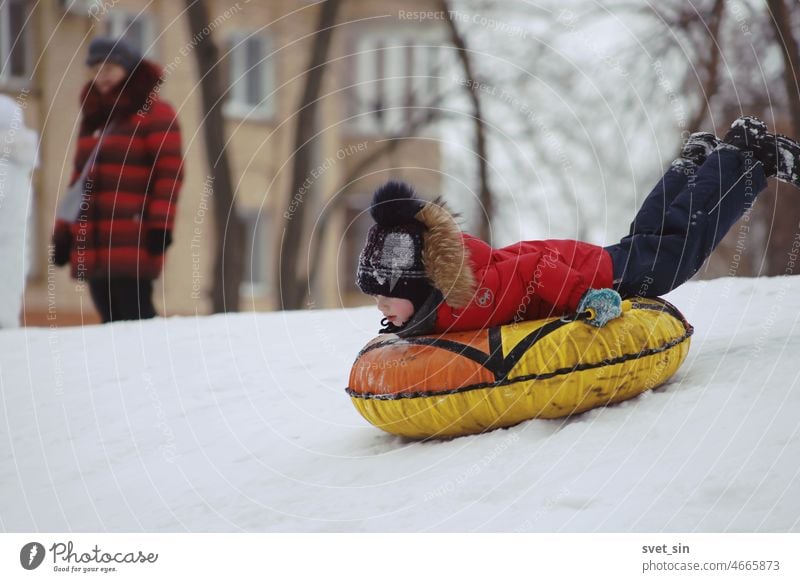 A boy on a bright tubing slides down a snowy hill against the backdrop of the urban landscape. Boy (child) Winter Snow Snowy hill roll down fun season Child