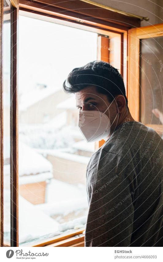 Young man with mask in quarantine looking at the snowfall through the window of his house alone anxiety boy confined confinement contemplation corona