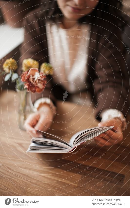 Woman reading a book in cafe sitting at wooden table with flowers in a vase. Romantic date with a book. Brown vintage neutral color background. Literature & Library, book lover spending leisure time.