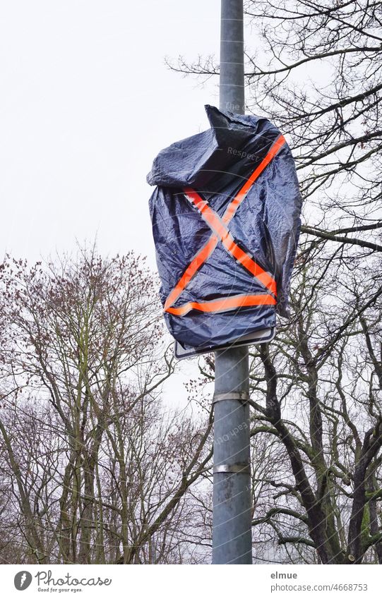 Traffic sign and additional sign, covered with a blue plastic bag and orange adhesive strips in front of bare trees / road traffic / construction measure