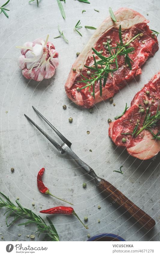 Close up of raw beef steak with rosemary and green pepper, garlic and butcher fork close up chili concrete kitchen table preparing fresh meat homemade bbq