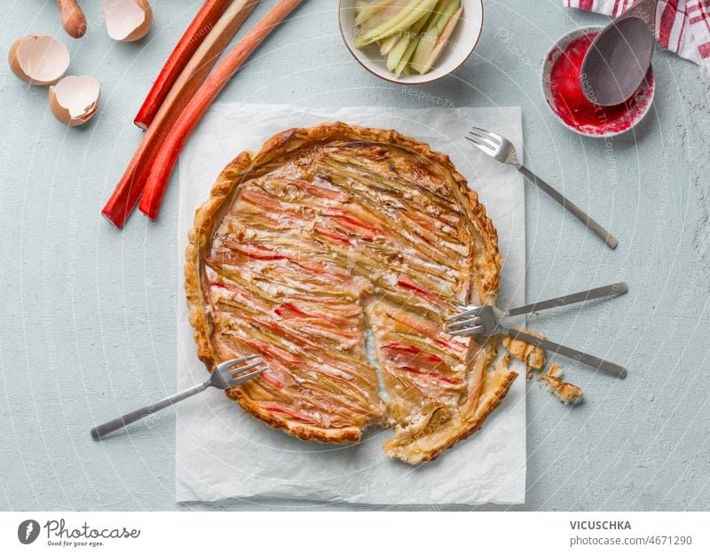 Homemade rhubarb cake at pale blue kitchen table with forks and ingredients homemade rhubarb sticks eggshells bowls seasonal summer top view baked delicious