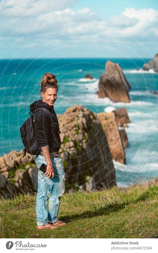 Smiling woman standing on coast with blue seawater traveler hill cliff wanderlust journey freedom positive explore female ocean nature smartphone trip tourism