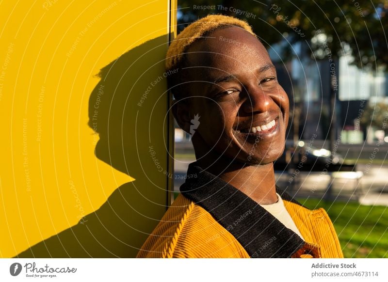 Joyful black man near wall on street style trendy urban appearance sunlight city dyed hair modern young casual short hair town male positive happy smile delight