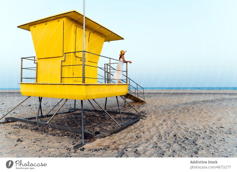 Woman standing near lifeguard hut woman beach shore alone contemplate travel sand coast yellow tower building sea female observe seashore seaside cabin color