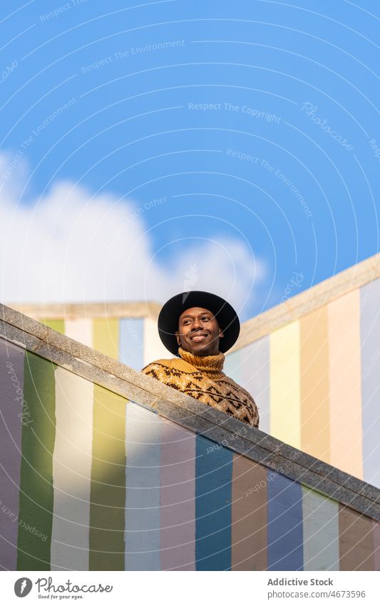 Black man in warm sweater on steps of staircase against striped colorful wall warm clothes cap trendy knitted casual portrait fashion male outfit style