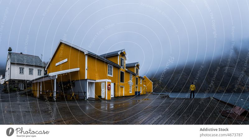 Anonymous tourist standing on pier near yellow house and admiring wild nature man admire mountain lake traveler fjord fog landscape wanderlust recreation male