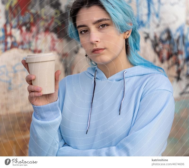 Blue haired Teenage girl in light blue oversize hoodie staying near graffiti wall with coffee to go Teenager mockup cup jeans blue haired teen girl outdoors