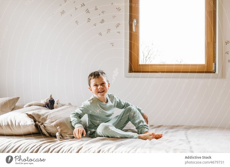 Cheerful boy sitting on bed kid window bedroom pajama childhood domestic morning having fun funny apartment adorable flat cute home residential nightwear dwell