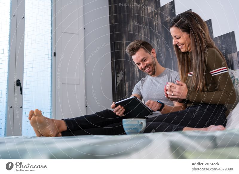 Cheerful couple browsing tablet on bed bedroom internet online coffee pastime relationship hobby free time home read sofa together device apartment gadget