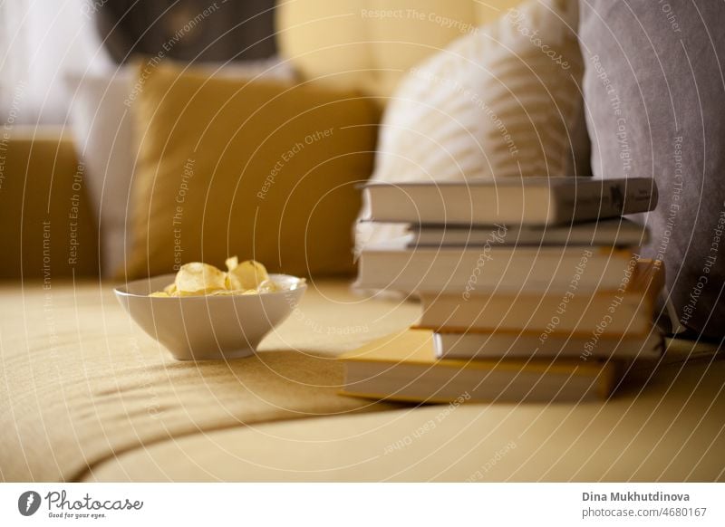 A stack of books and potato chips on cozy  yellow couch with pillows. Apartment living. Simple life. Millennial casual lifestyle. Staycation. Education. tasty