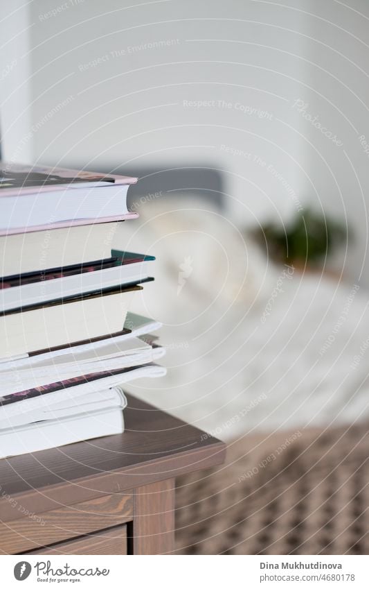 A stack of books on the woodenchest of drawers near cozy bed in the bedroom with neutral colors interior design. Cosy apartment living and reading background.