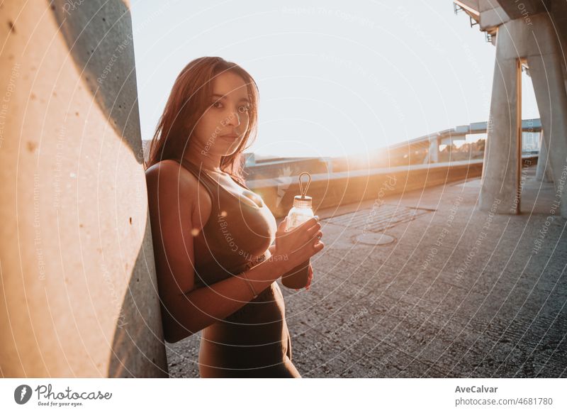 Young woman silhouette resting against a wall holding a bottle of water during a colorful sunset with copy space. Resting after workout on urban street ambient concept. Moroccan girl