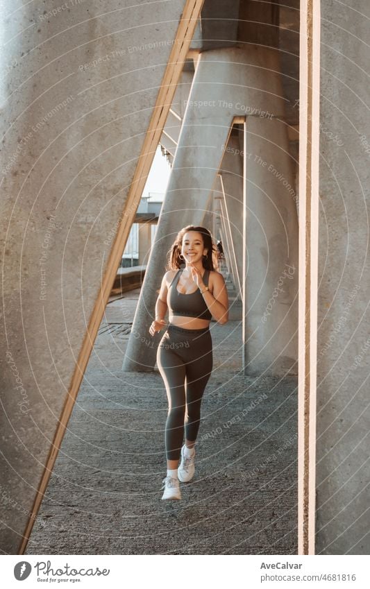 Young multi cultural woman smiling to camera while jogging on urban city to lose weight before summer to get a defined body. Running and workout outdoors. Sunset scene with young athletes