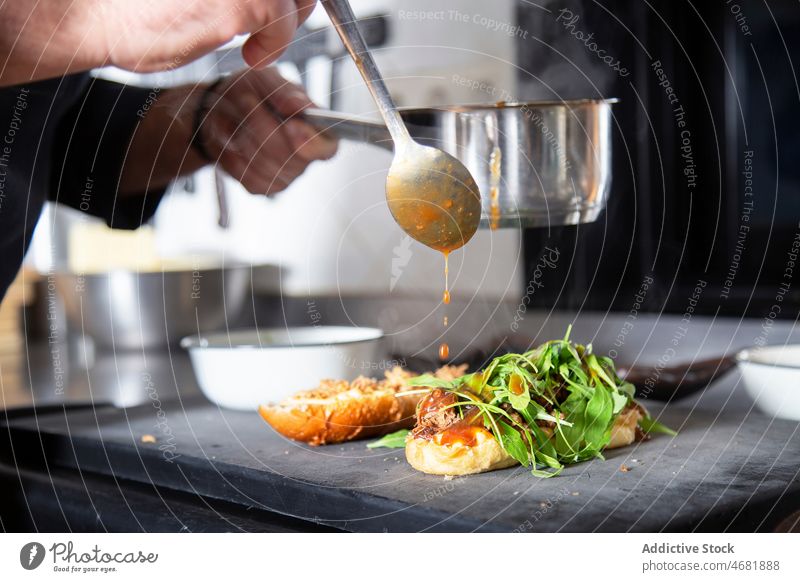 Unrecognizable chef pouring sauce on sandwich man bread cook kitchen cuisine culinary recipe meat greens herb saucepan restaurant work prepare counter job