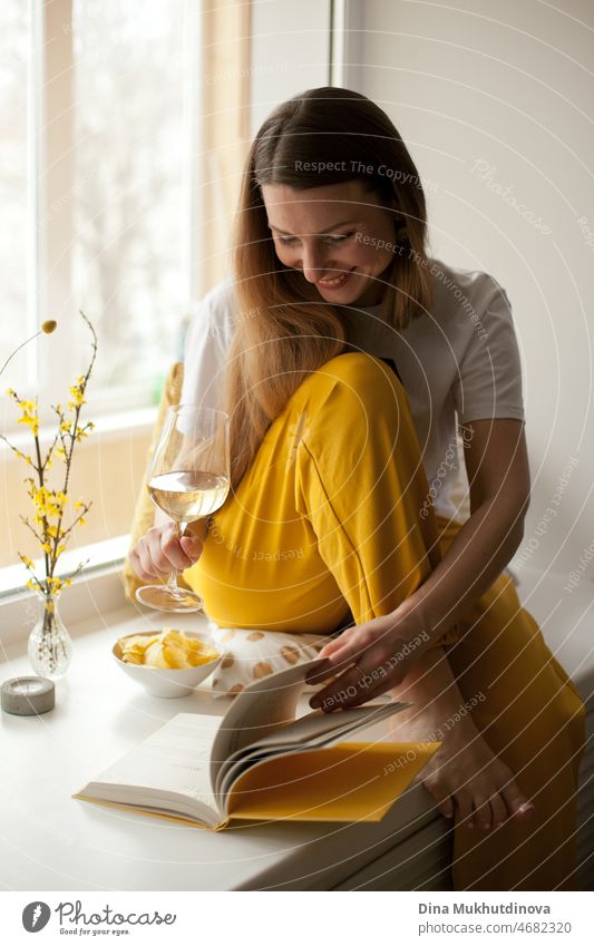 Young beautiful woman wearing gray and yellow sweatpants reading a book at home, sitting on windowsill, smiling and drinking white wine. Education and knowledge. Literature and fiction books.  Cozy home room.