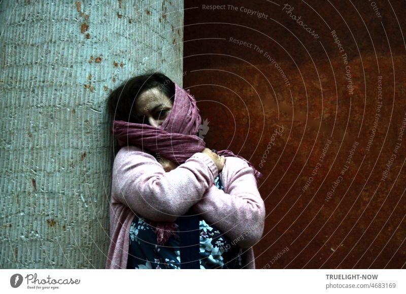 A mysterious woman in light purple cardigan over blue and white patterned dress partially veils her arms crossed in front of her chest her face and dark hair with a mauve taupe colored scarf in front of rust and mint colored old walls