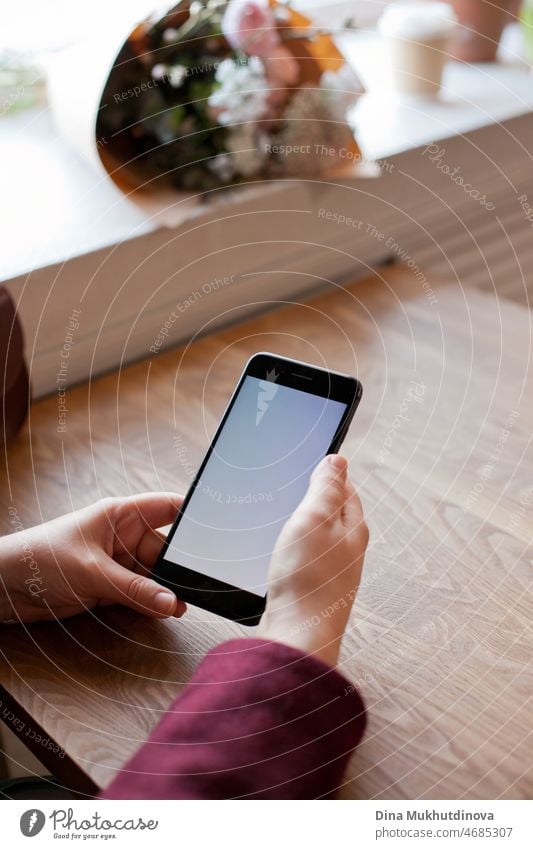 Woman hands holding a mobile phone texting close-up mockup. Blank screen of a smartphone on wooden table background. Woman using technology at cafe. Empty screen of cellphone in female hands.