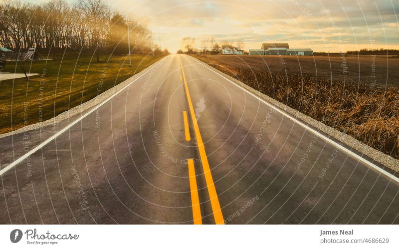 The sun rises over the rural country highway with a farm silhouetted on the horizon. Fields and trees follow the sides of the road. sunshine treelined vivid