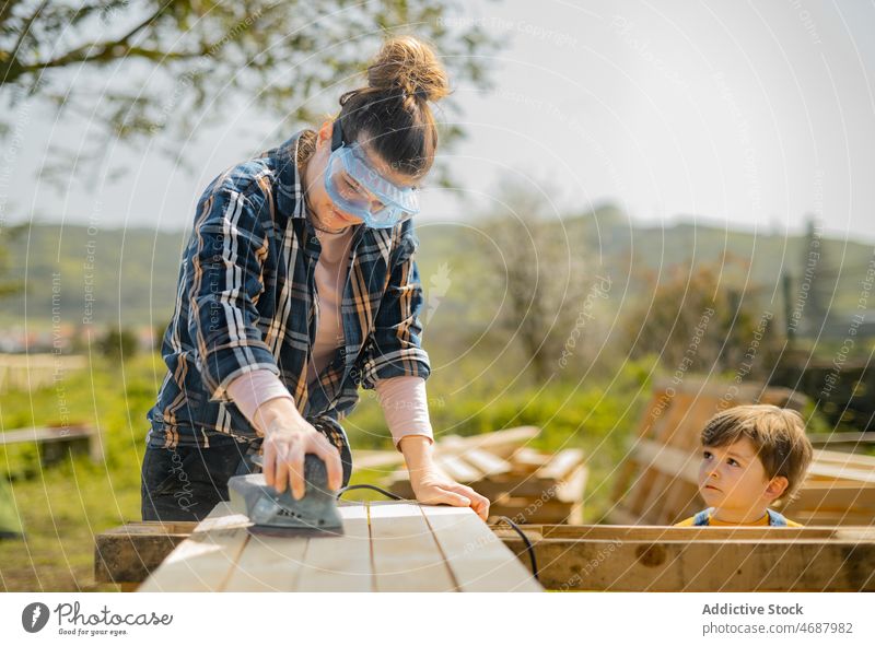 Woman sanding down wooden detail near boy woman sander instrument tool plank countryside board woodwork focus carpenter craftswoman electric summer female