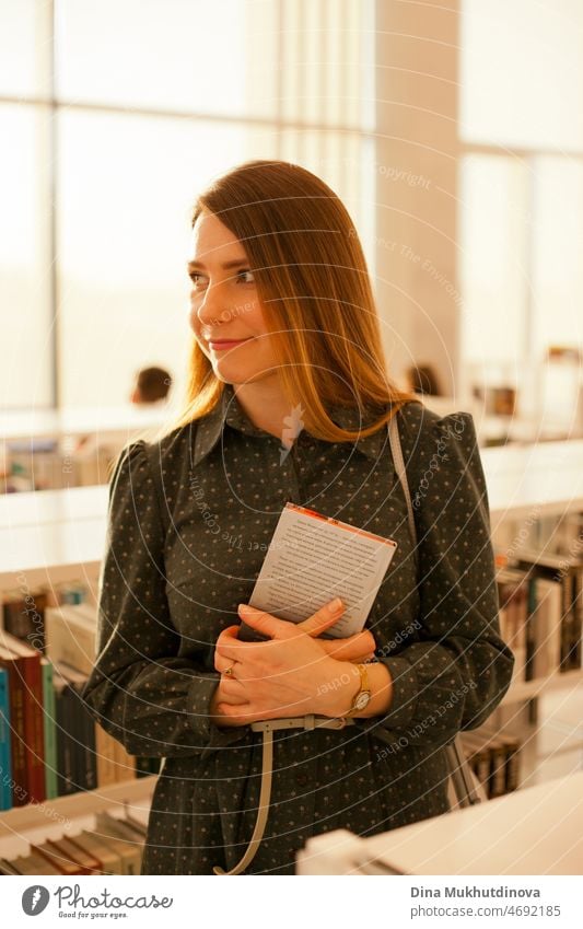 Adult female student at university library holding a book. Beautiful woman at public library with a book standing between bookshelves. Candid lifestyle portrait. Books and education. Knowledge. College and university lifestyle.