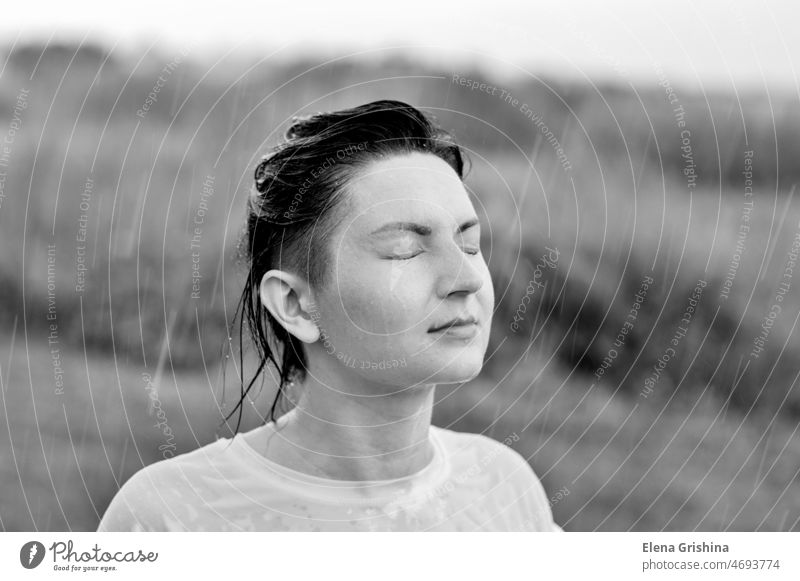 A  young girl stands with her eyes closed in the summer rain. wet white shirt wet clothes drops happy closed eyes wet hair brunette natural background day