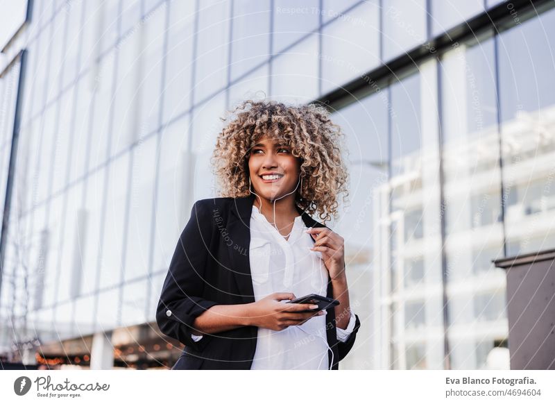 beautiful smiling business woman using mobile phone and headphones in city. Buildings background afro hispanic skyscraper building young curly hair music