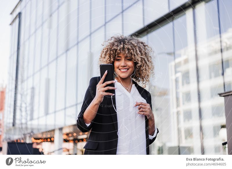 beautiful smiling business woman using mobile phone and headphones in city. Buildings background afro hispanic skyscraper building young curly hair music